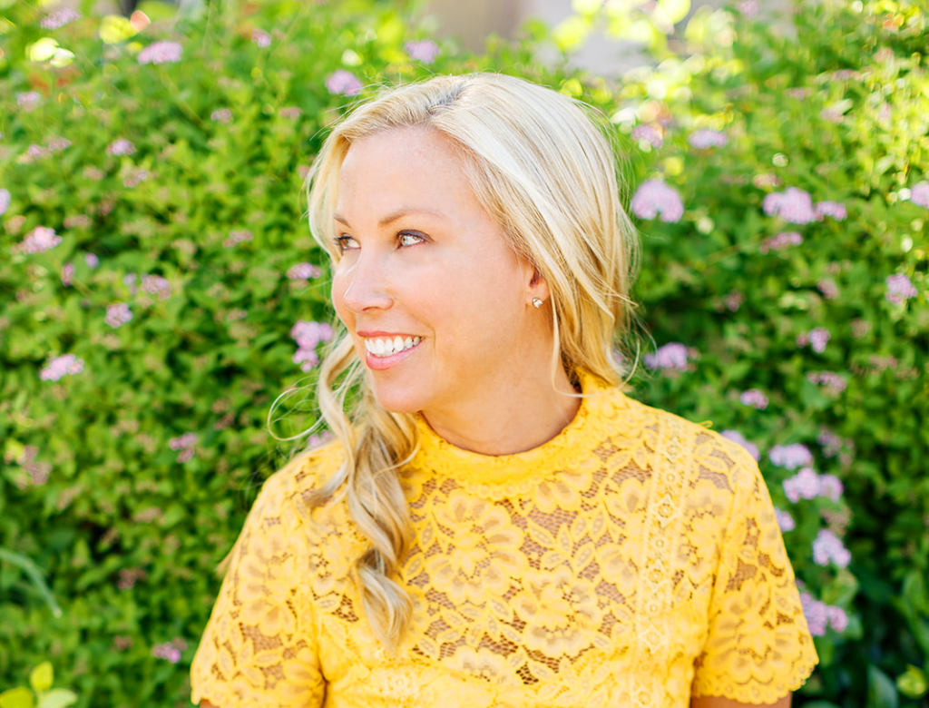Woman is wearing a bright yellow top and is smiling and is surrounded by flowers